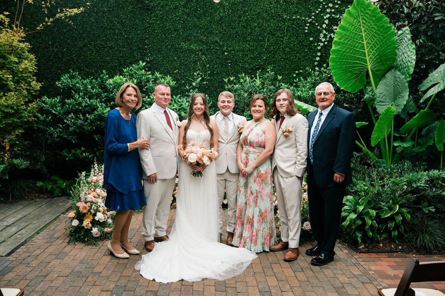 Wedding party in light summer attire poses against lush green garden backdrop.