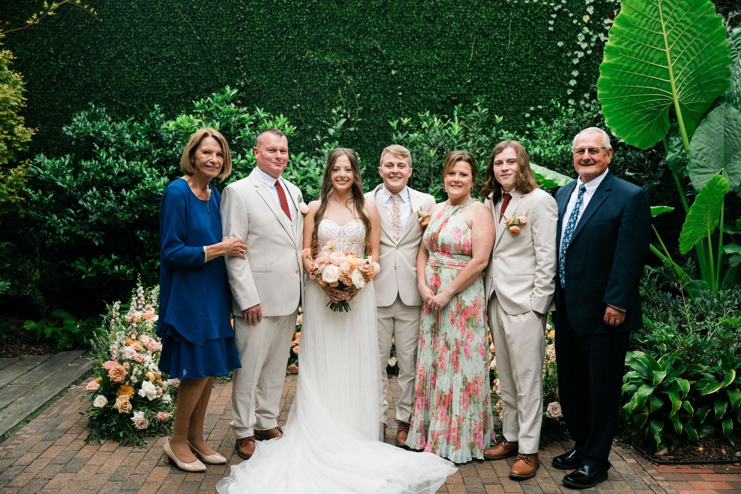 Wedding party poses for a formal group photo in front of a lush garden backdrop.