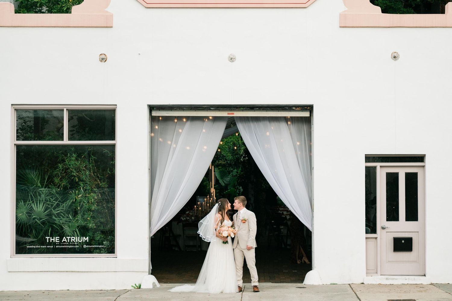 A couple in white wedding attire stands in front of a building entrance with flowing white curtains and greenery.