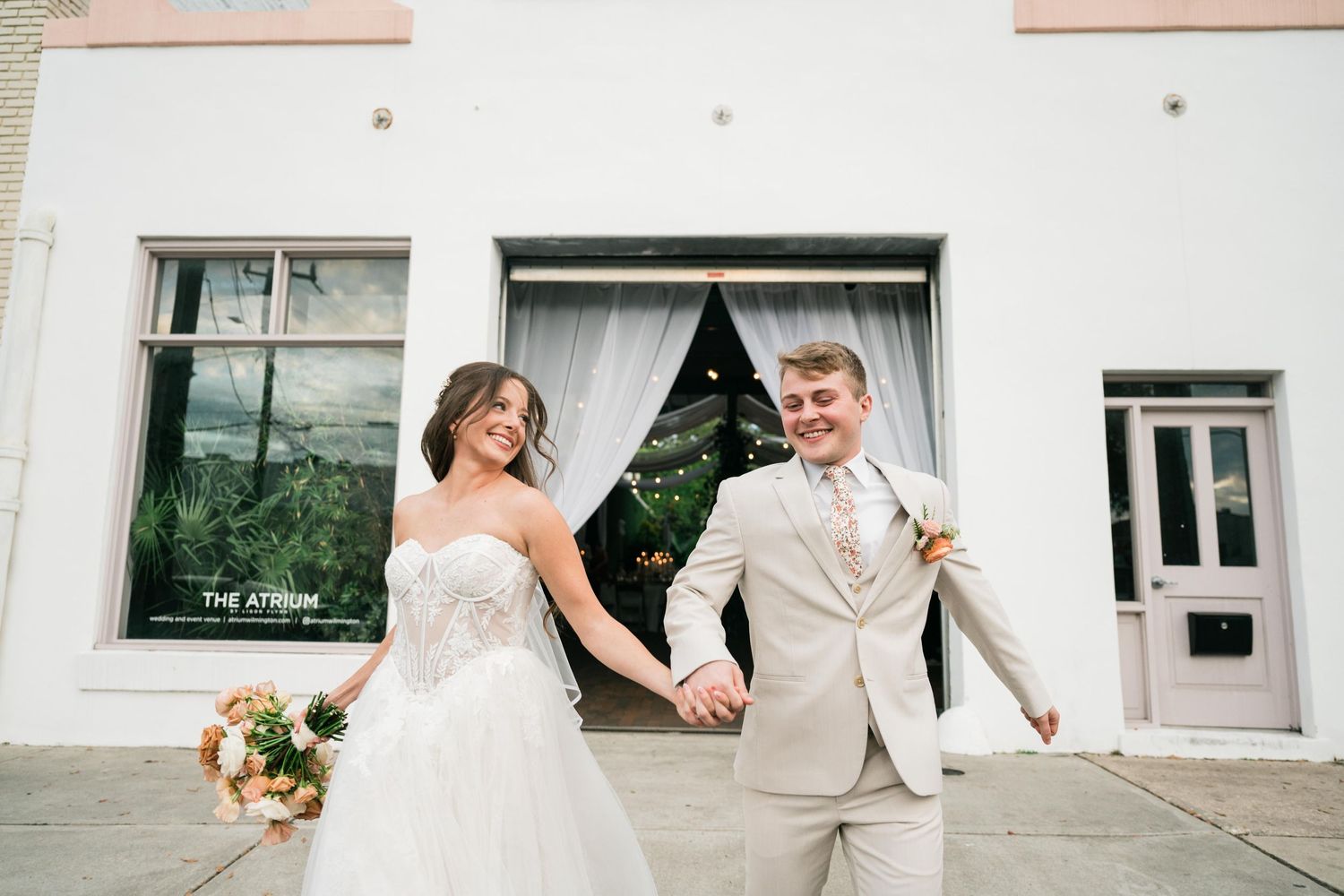 A joyful couple in formal wedding attire holds hands while walking along a sidewalk by a white building.