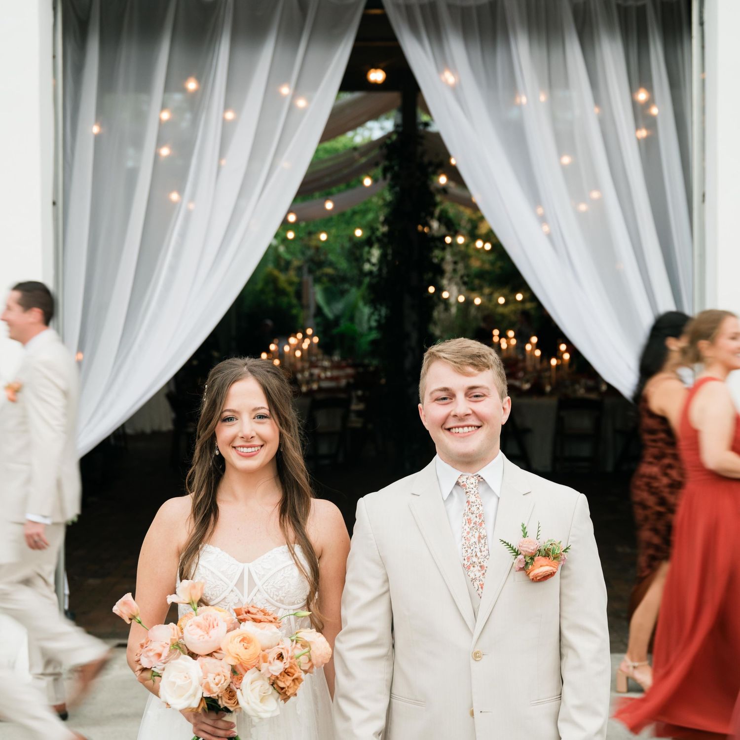 Happy newlyweds stand together under white draped fabric and string lights at an outdoor wedding reception.