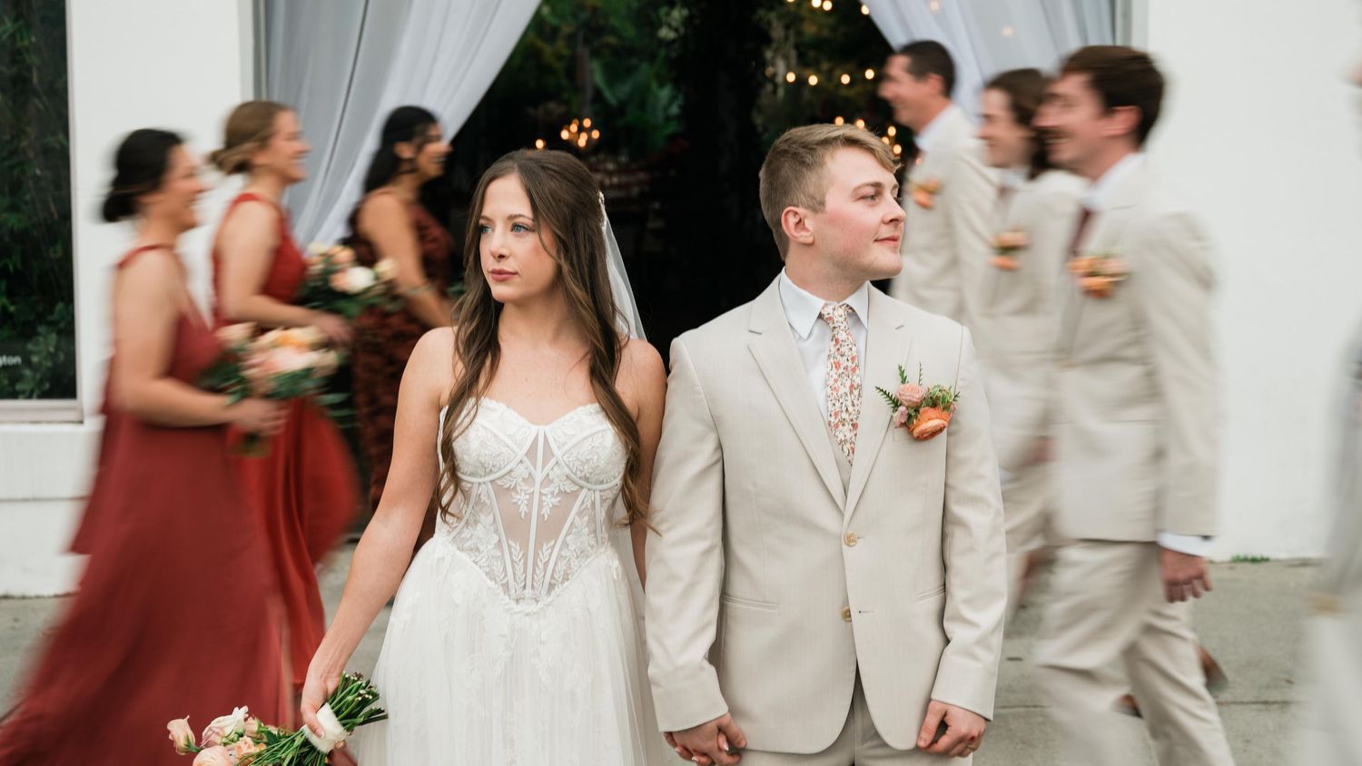 Wedding party in formal attire with bridesmaids in red dresses and groomsmen in light beige suits during outdoor ceremony.