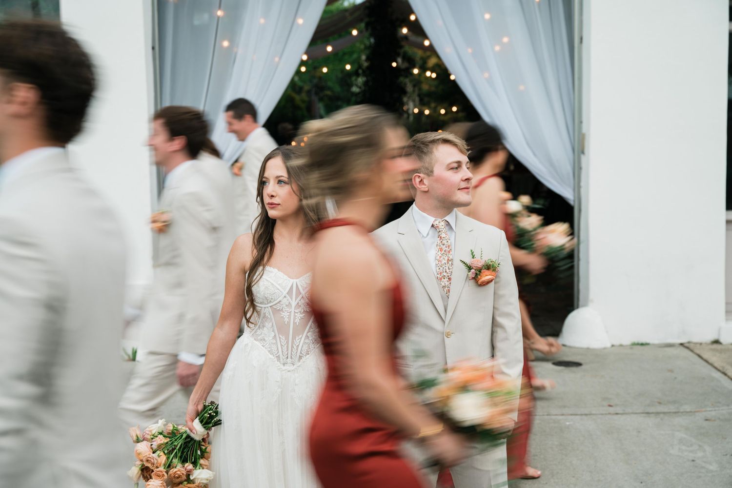 Blurred motion captures wedding party members in light colored formal attire walking past white curtains and a Christmas tree.