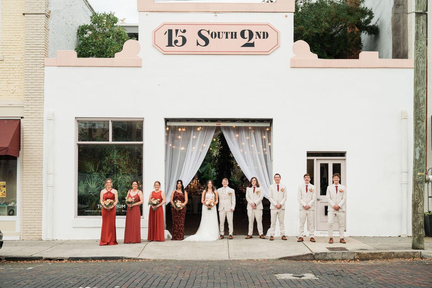 A wedding party poses in front of a charming brick storefront with white curtains and red and neutral attire.