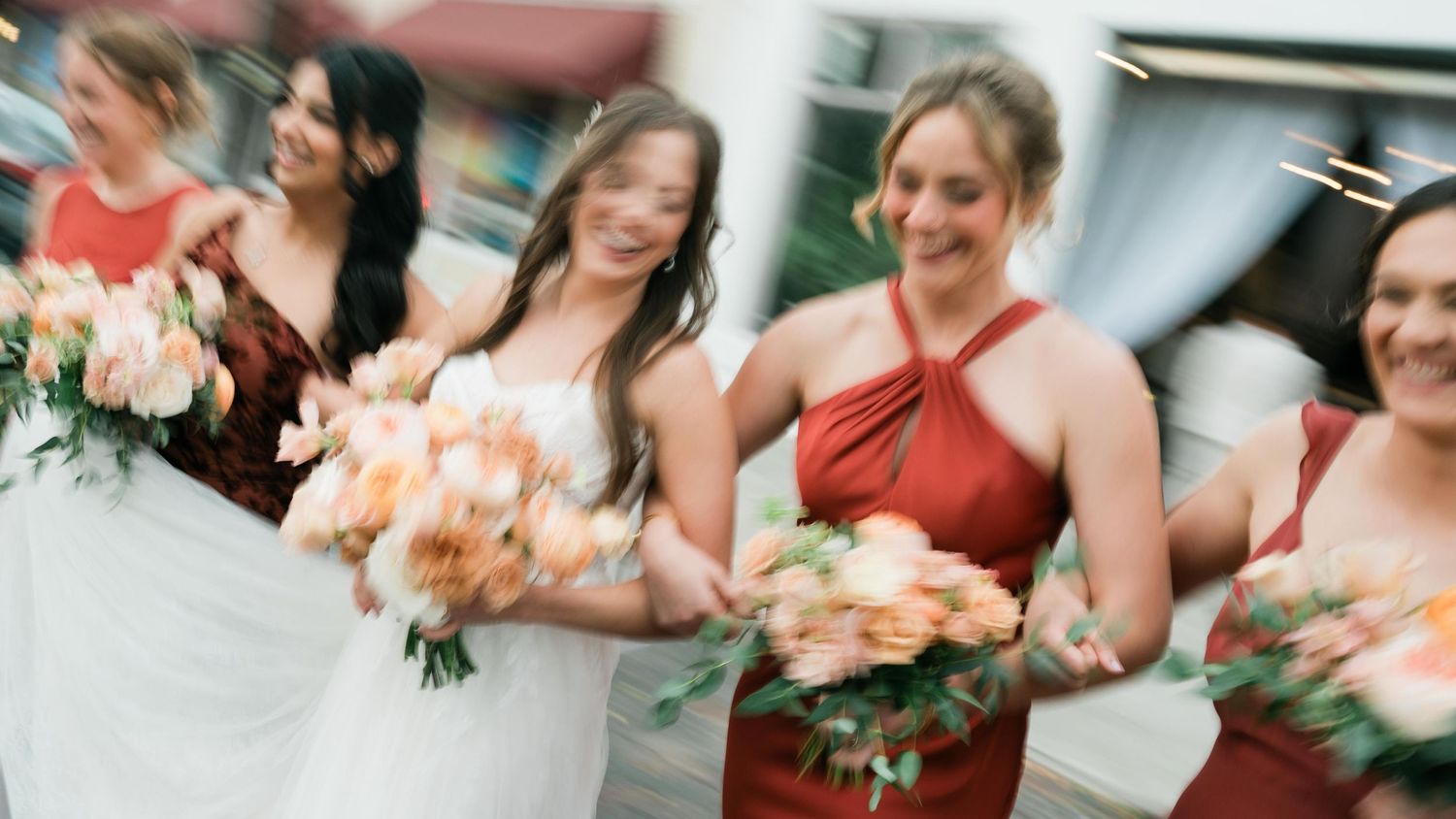 A candid moment captures a bride with bridesmaids holding peach and coral colored bouquets at an outdoor wedding.