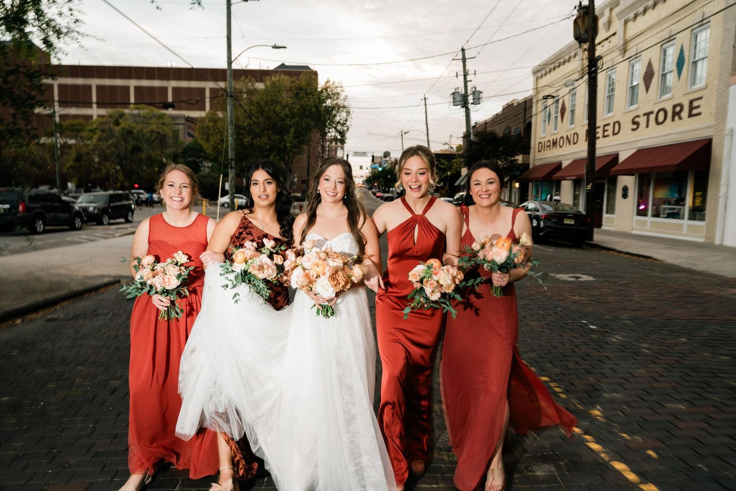 Wedding party in rust-colored dresses walks down cobblestone street with bouquets and bridal gown.