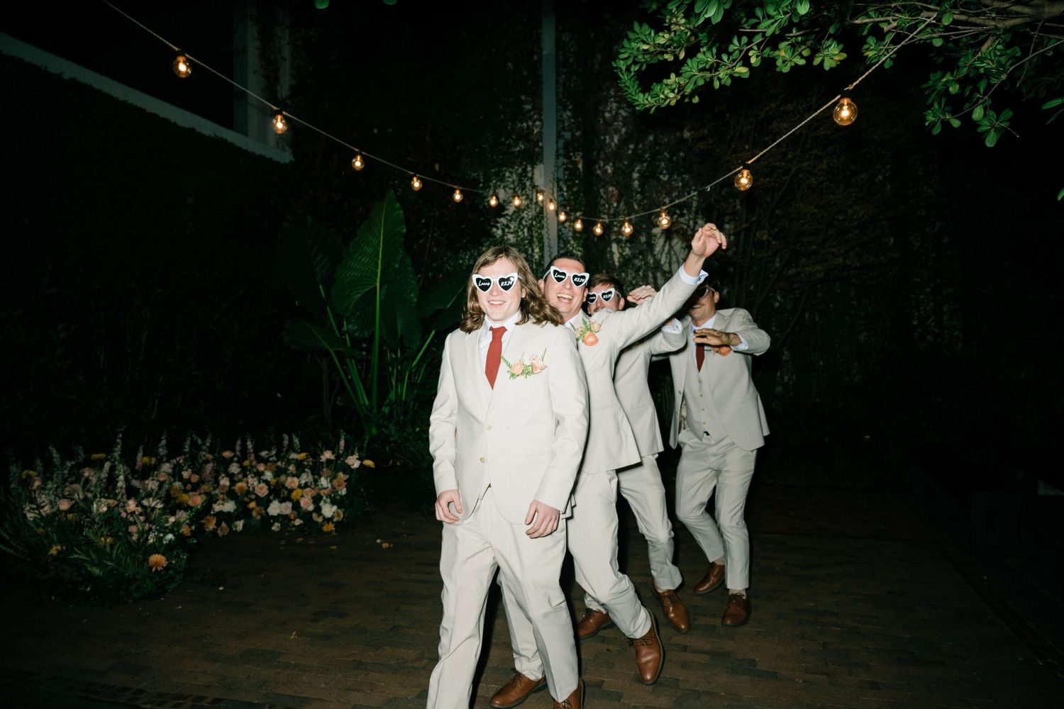 Wedding party members joyfully dancing and jumping in white and gray suits at an evening outdoor reception.