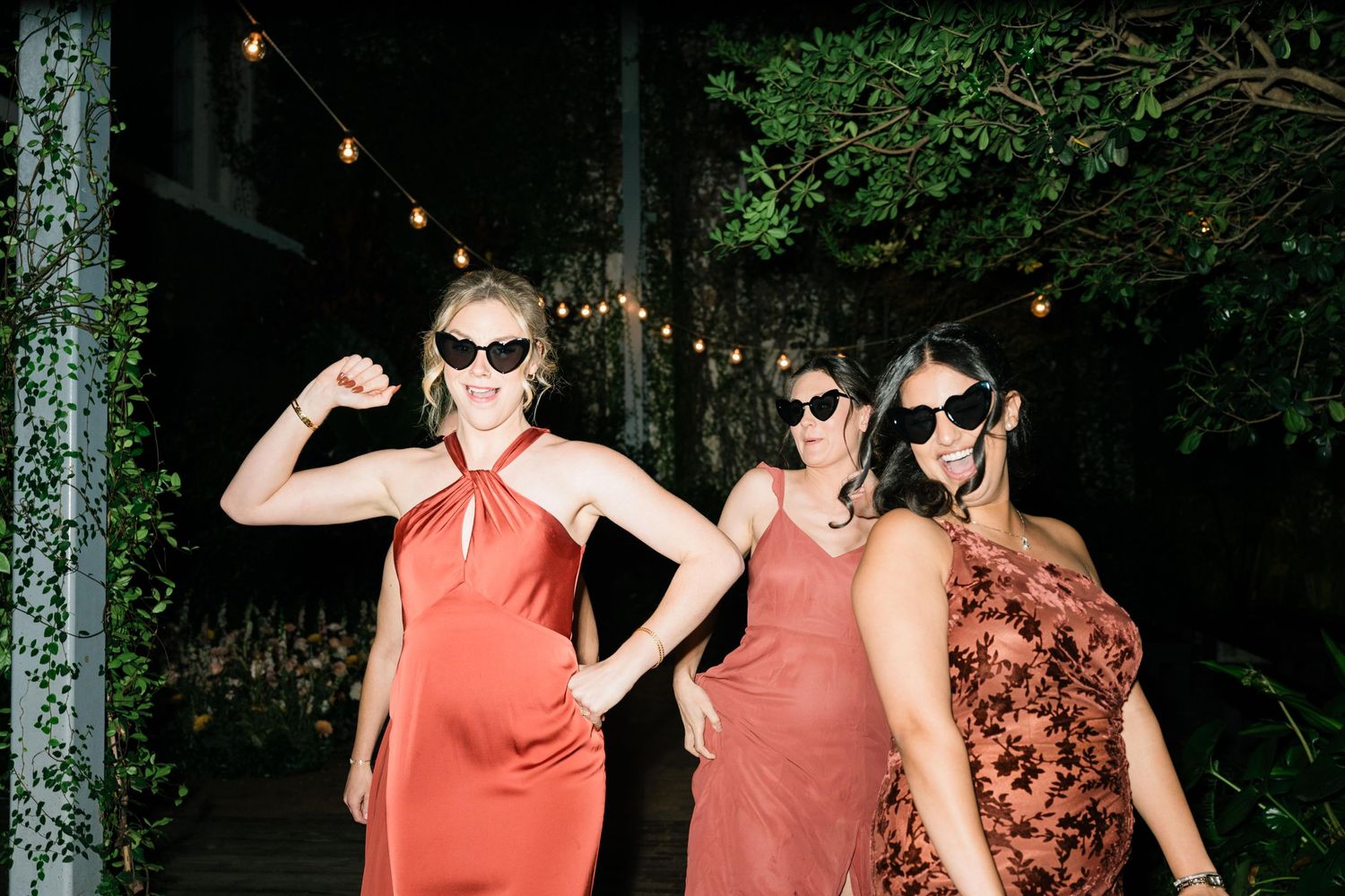 Three women in coral dresses and sunglasses pose at a nighttime outdoor party under string lights and trees.