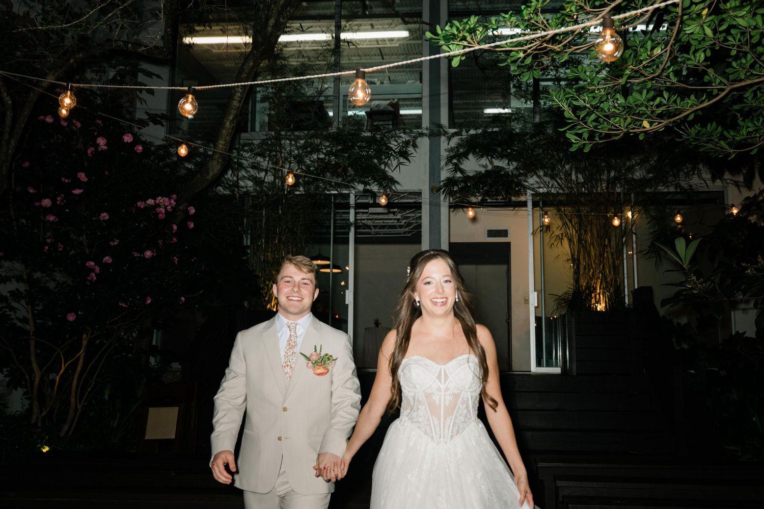 A couple walks hand in hand at a nighttime wedding reception under string lights and greenery.