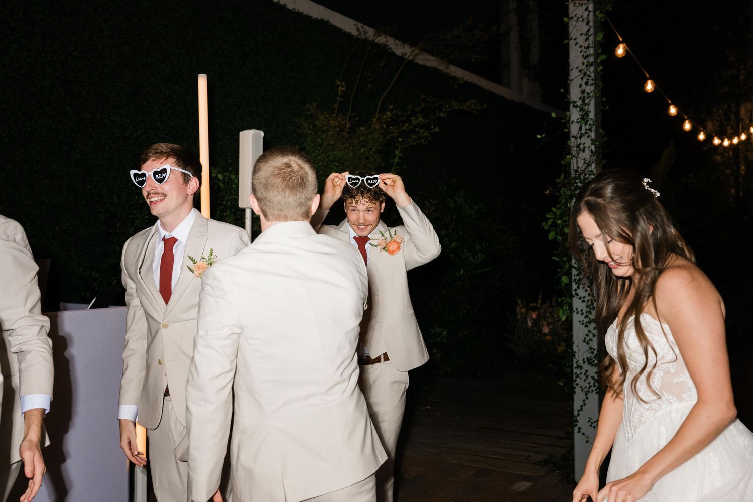 People in light colored formal attire celebrating at an outdoor evening reception with string lights overhead.