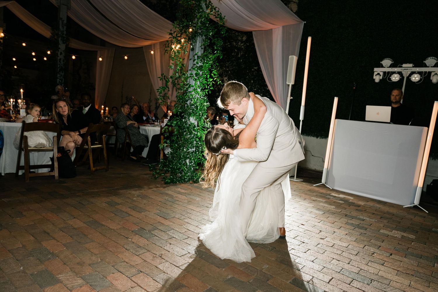 Couple shares romantic dip kiss on wooden dance floor under twinkling lights at outdoor reception.