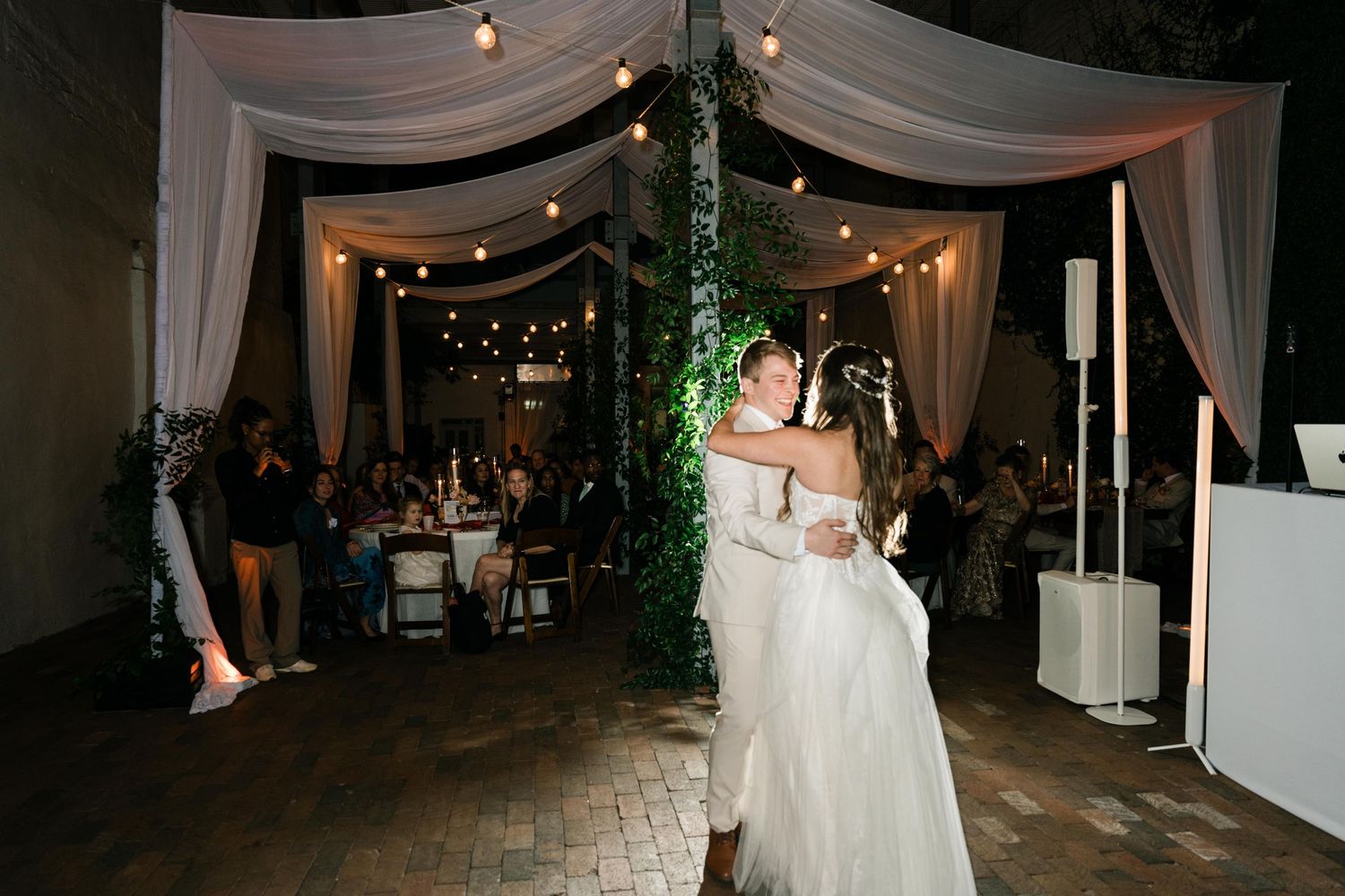 A romantic wedding reception dance scene under a draped white canopy with twinkling lights and ivy-covered pillars.