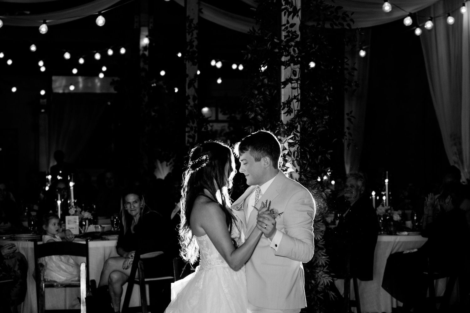 Black and white photo of newlyweds sharing their first dance under romantic string lights.