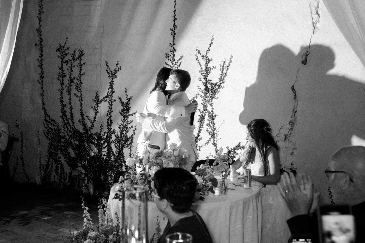 Black and white photograph of a wedding reception setup with shadows cast on a wall behind a table with floral arrangements.