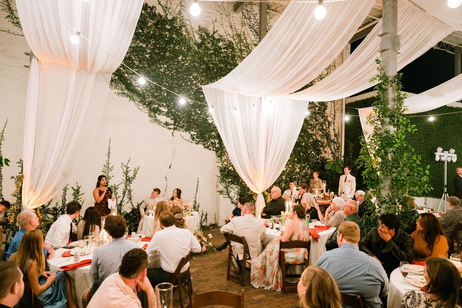 Wedding reception venue decorated with elegant white drapes and greenery as guests enjoy dinner.