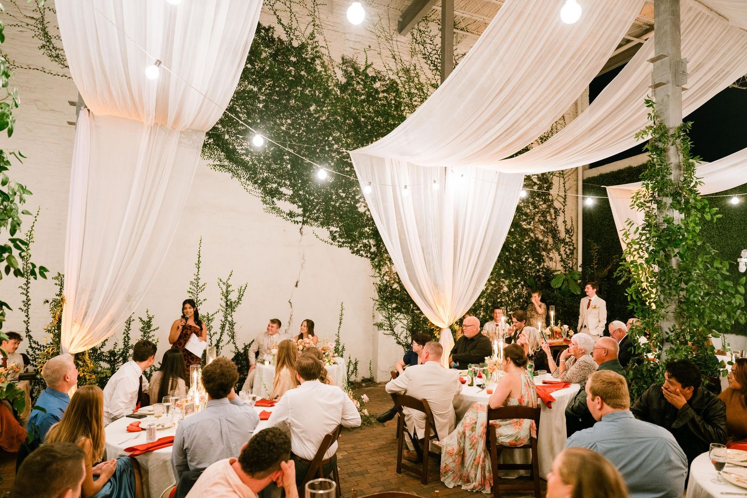 Indoor wedding reception venue decorated with flowing white drapes and greenery features guests dining at decorated tables.
