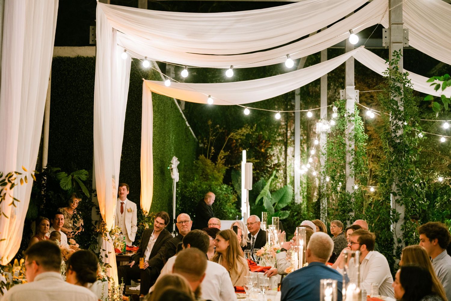 Elegant outdoor wedding reception under white fabric draping with hanging lights creating a romantic evening atmosphere.