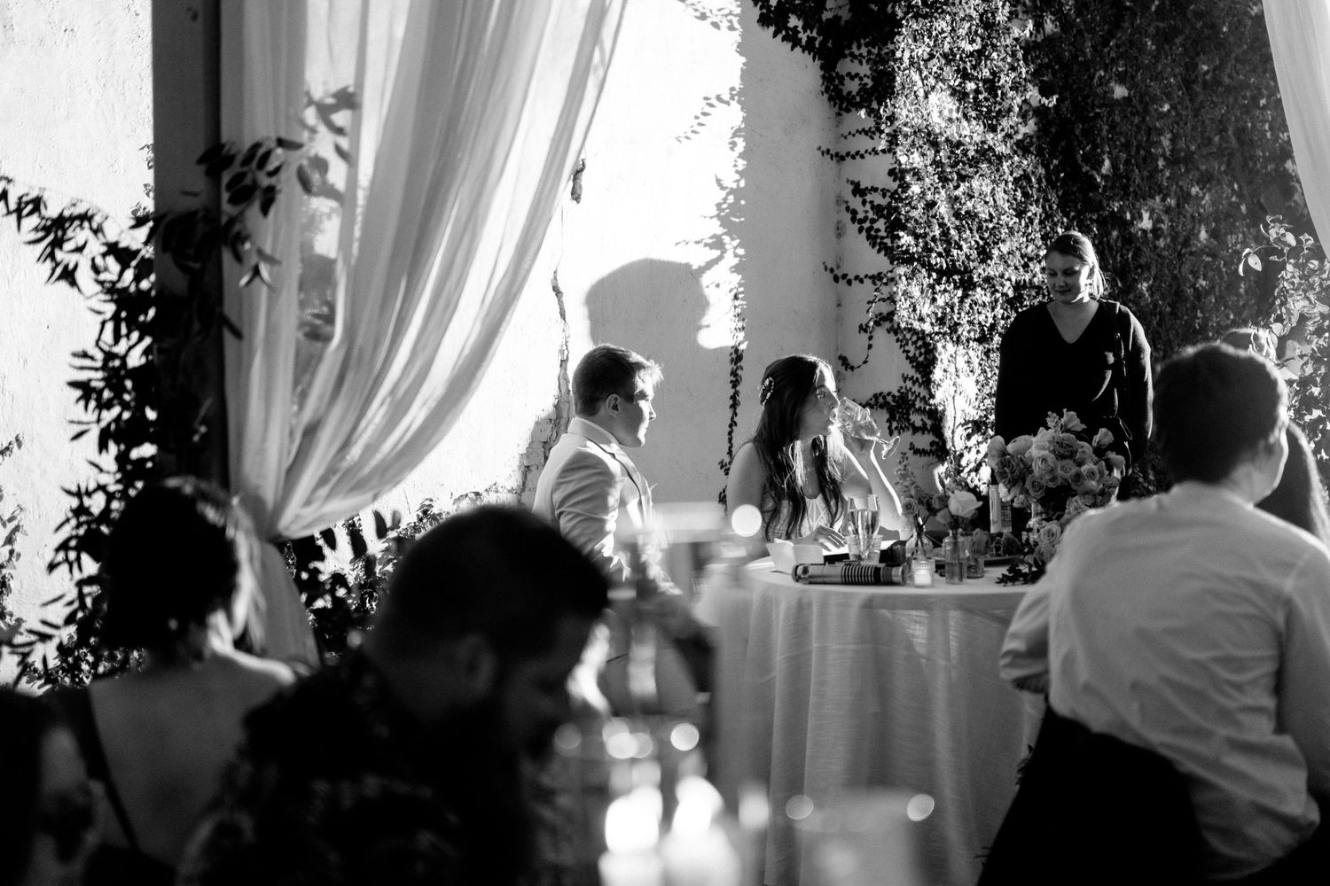 A candid black and white reception moment captured under elegant white drapes and foliage decor.