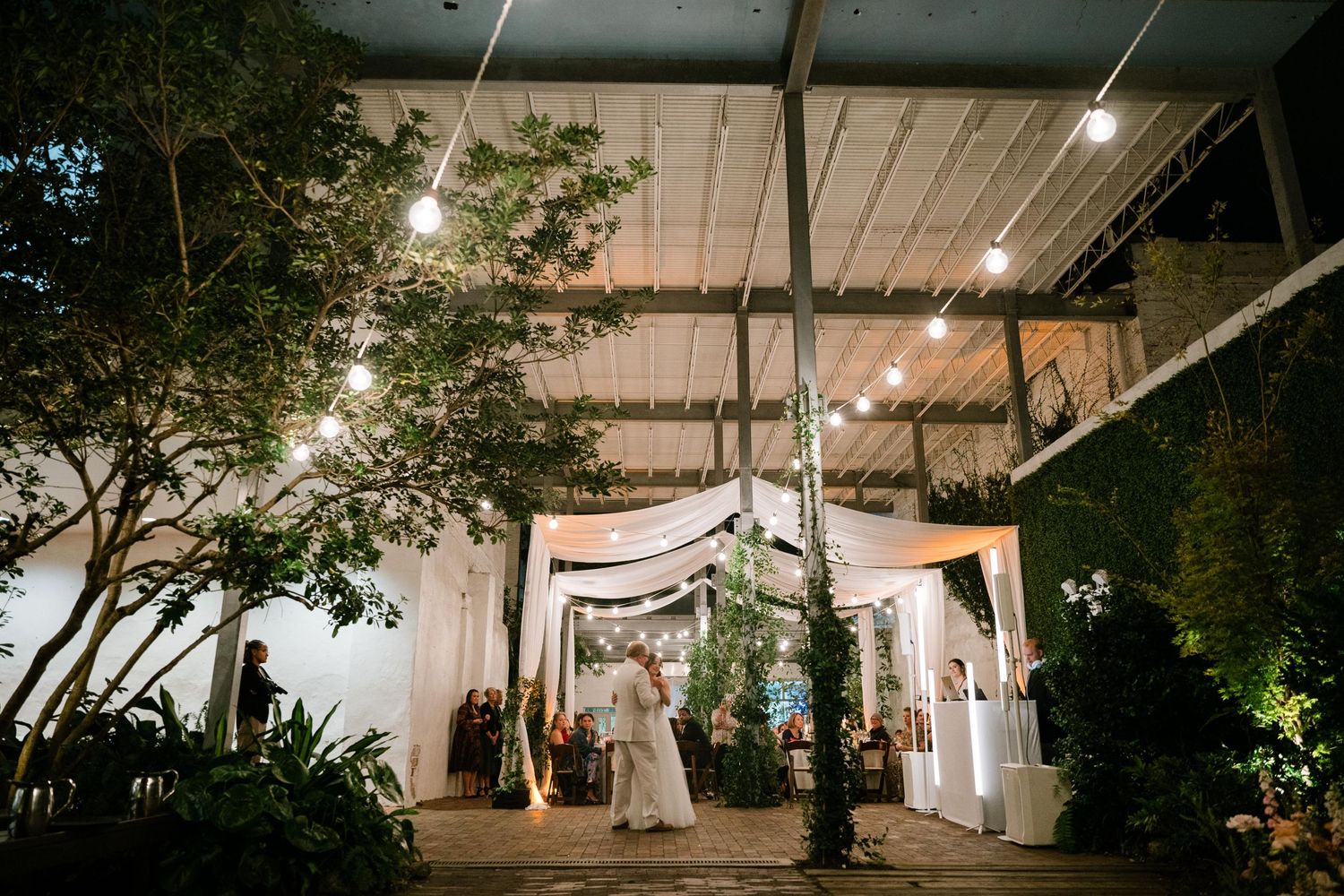 Couple enjoys an outdoor wedding reception at night under a decorated venue entrance with string lights and greenery.