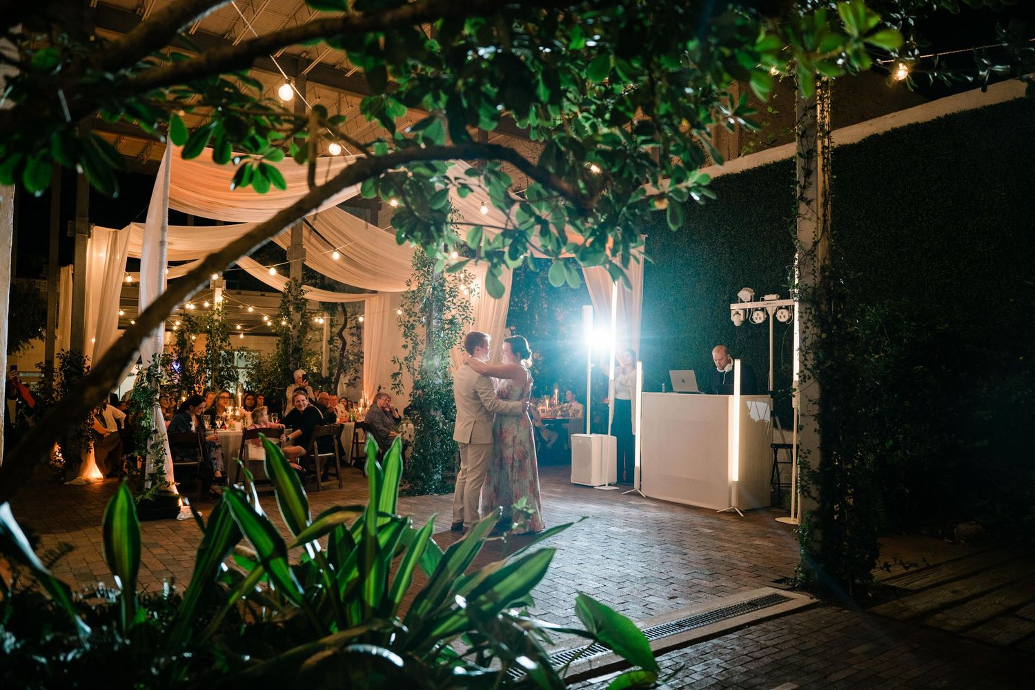 A romantic evening wedding reception scene with string lights, tropical plants and a DJ booth.