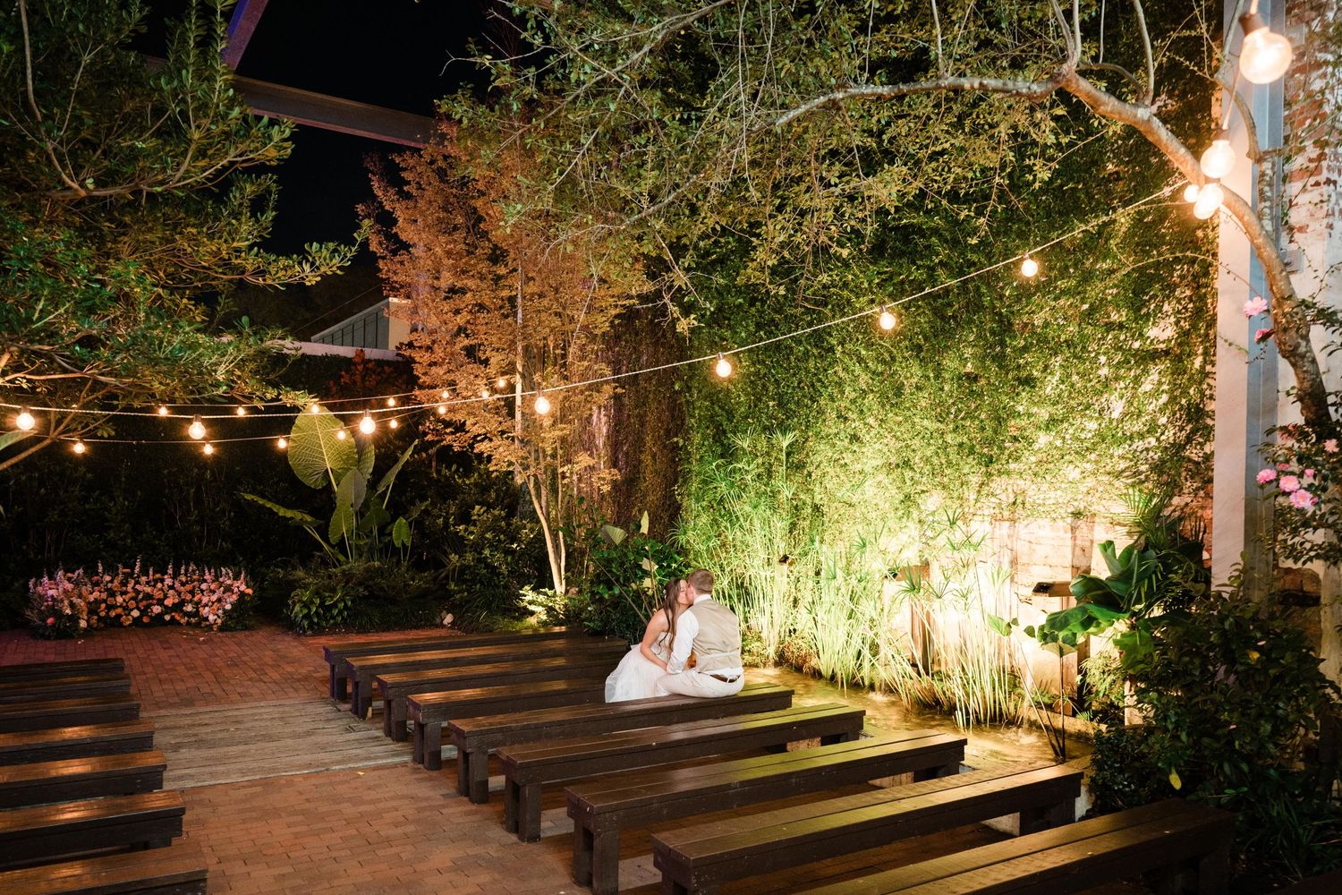 Outdoor wedding ceremony setup with wooden benches and string lights illuminating a garden at night.