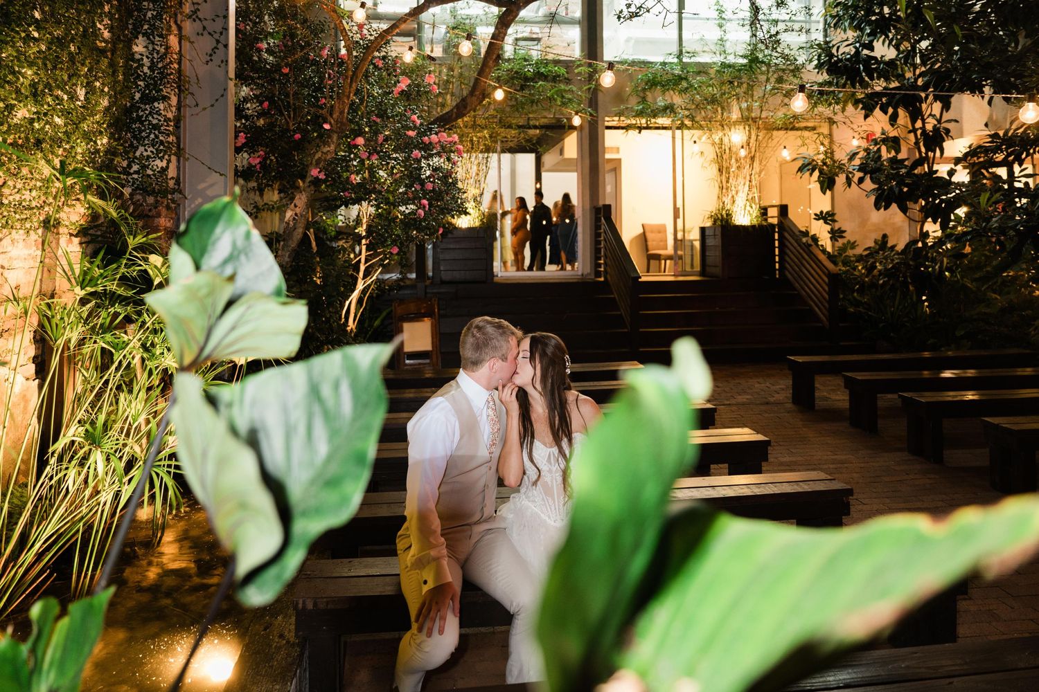 Two people share an intimate moment in a garden venue with large green leaves in the foreground and warm lighting.
