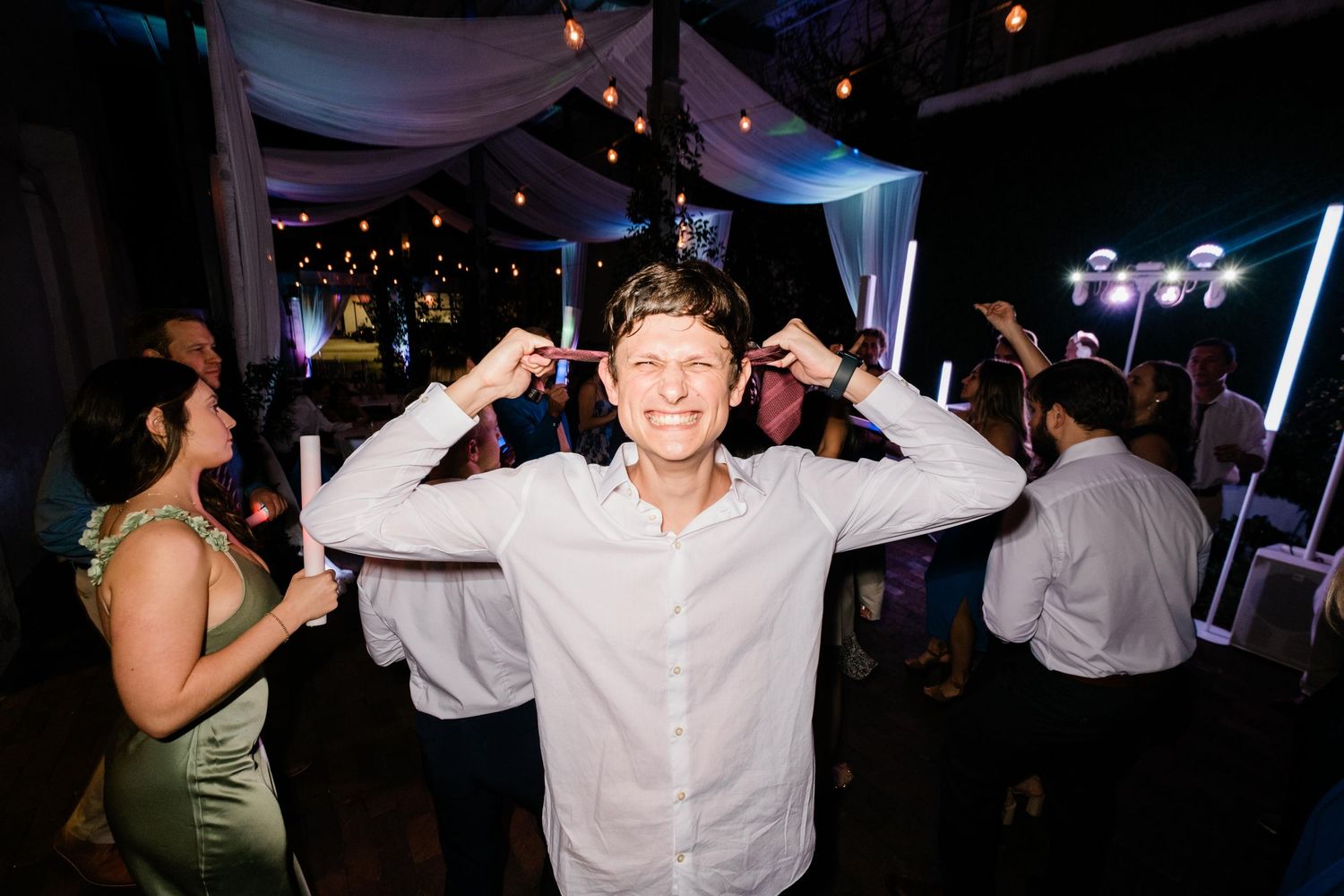 A joyful guest dances with raised arms at a wedding reception under draped fabric and twinkling lights.