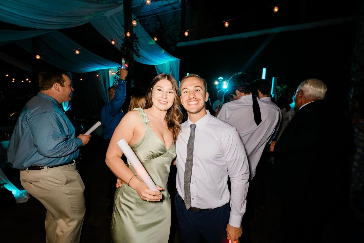 A couple celebrates at an evening reception with teal lighting and string lights decorating the venue.
