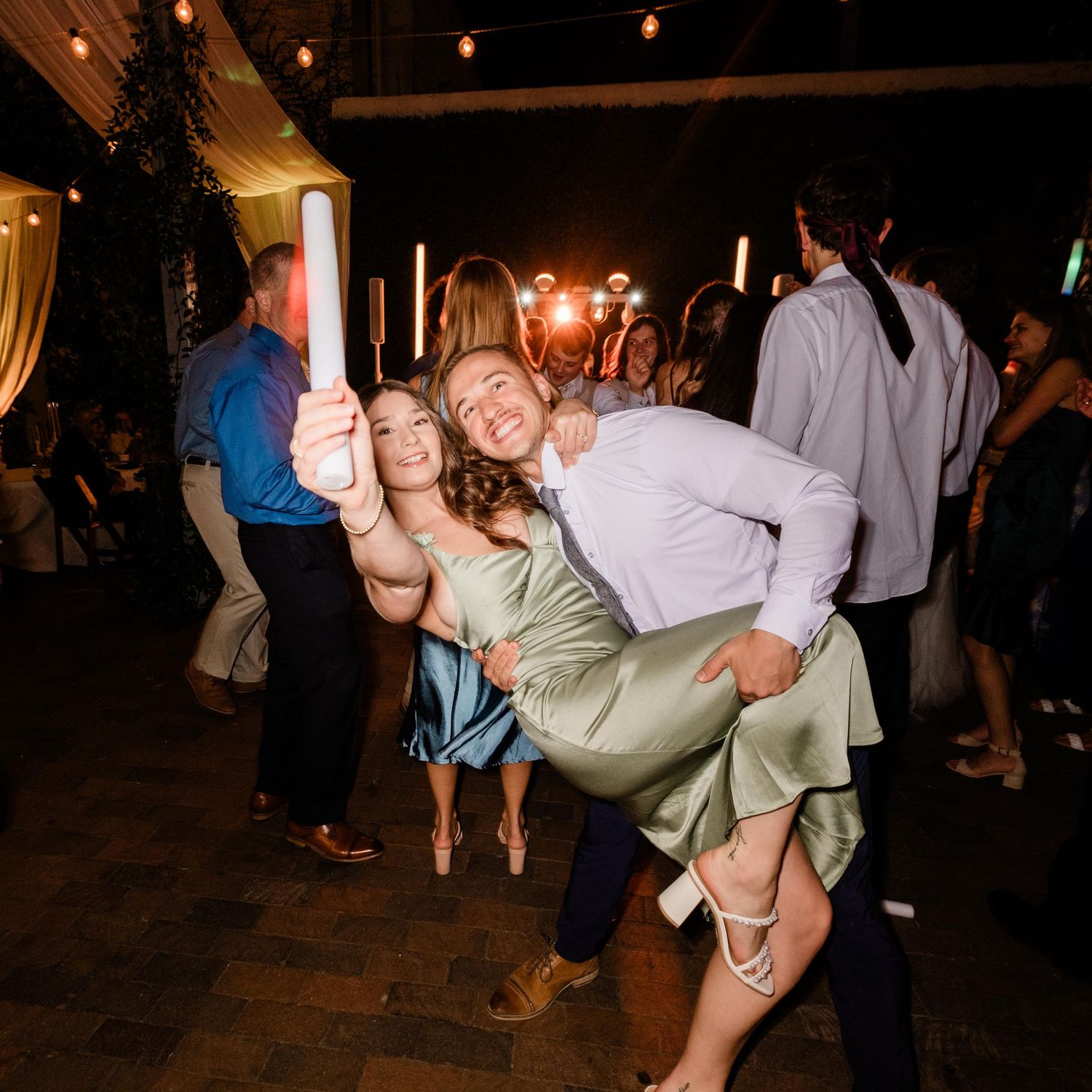 Wedding guests dancing energetically at a dimly lit reception with string lights overhead.