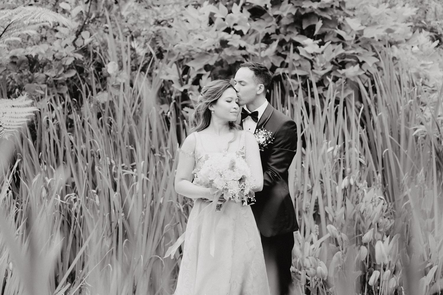 Romantic black and white wedding photo of a couple embracing among tall reeds and garden foliage.