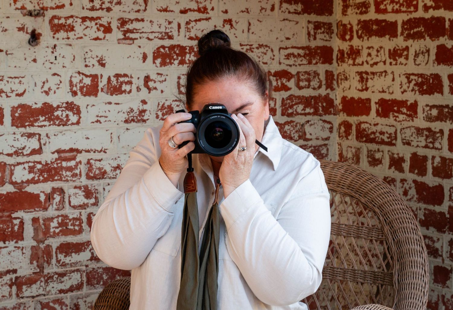 Photographer wearing a white shirt takes picture with professional camera against rustic brick wall background.