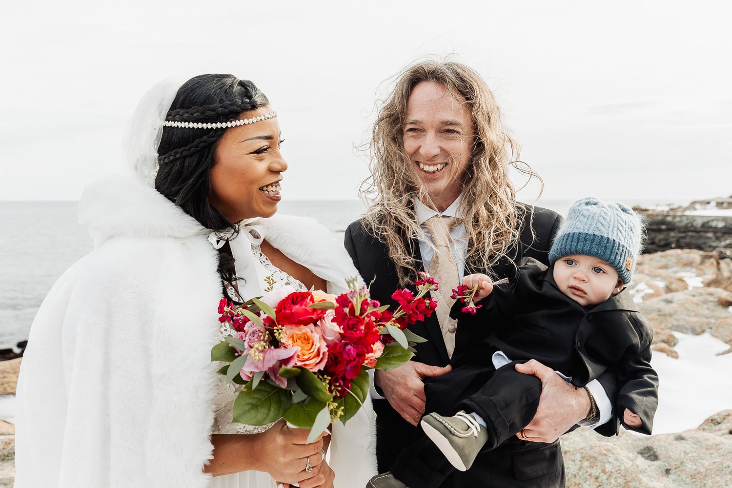 Three people share a joyful moment outdoors in winter, with one holding a colorful bouquet of red and pink flowers.