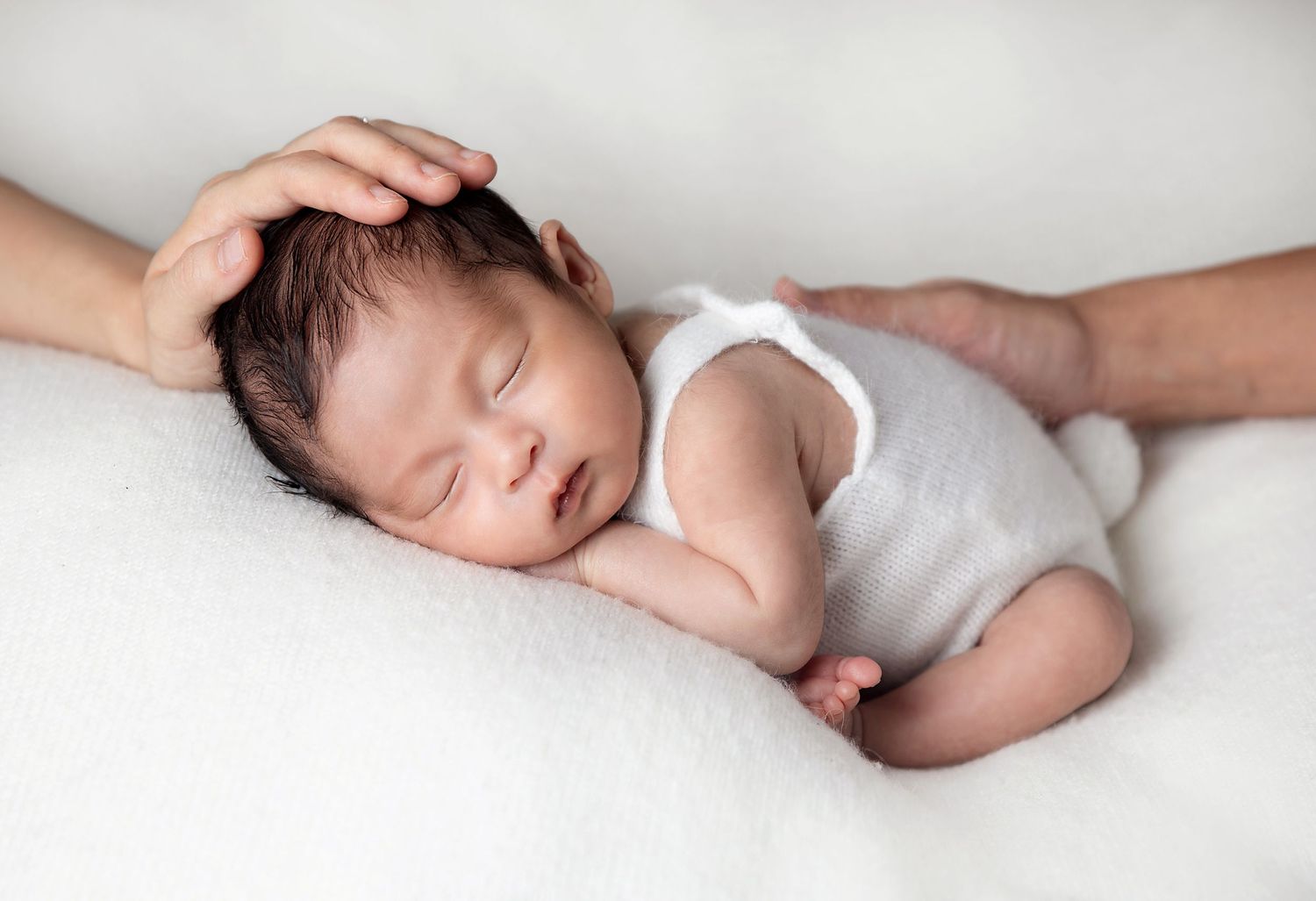 Peaceful sleeping newborn baby wearing grey onesie rests on soft white blanket while being gently cradled. During a photoshoot in Sidcup
