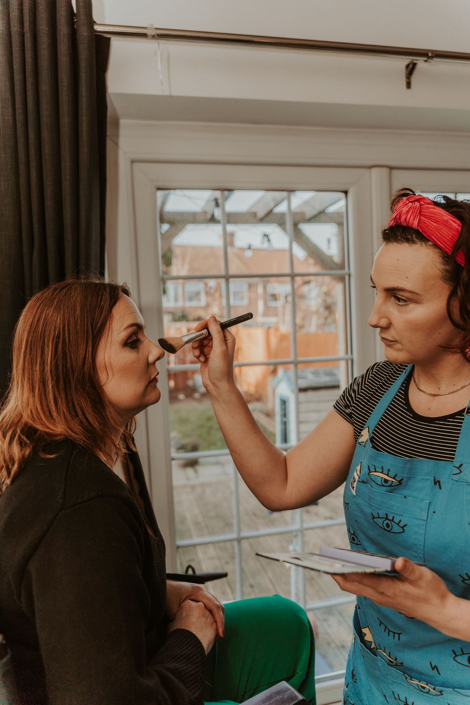 A makeup artist wearing blue overalls and red headband applies cosmetics near a window in a natural-lit room.