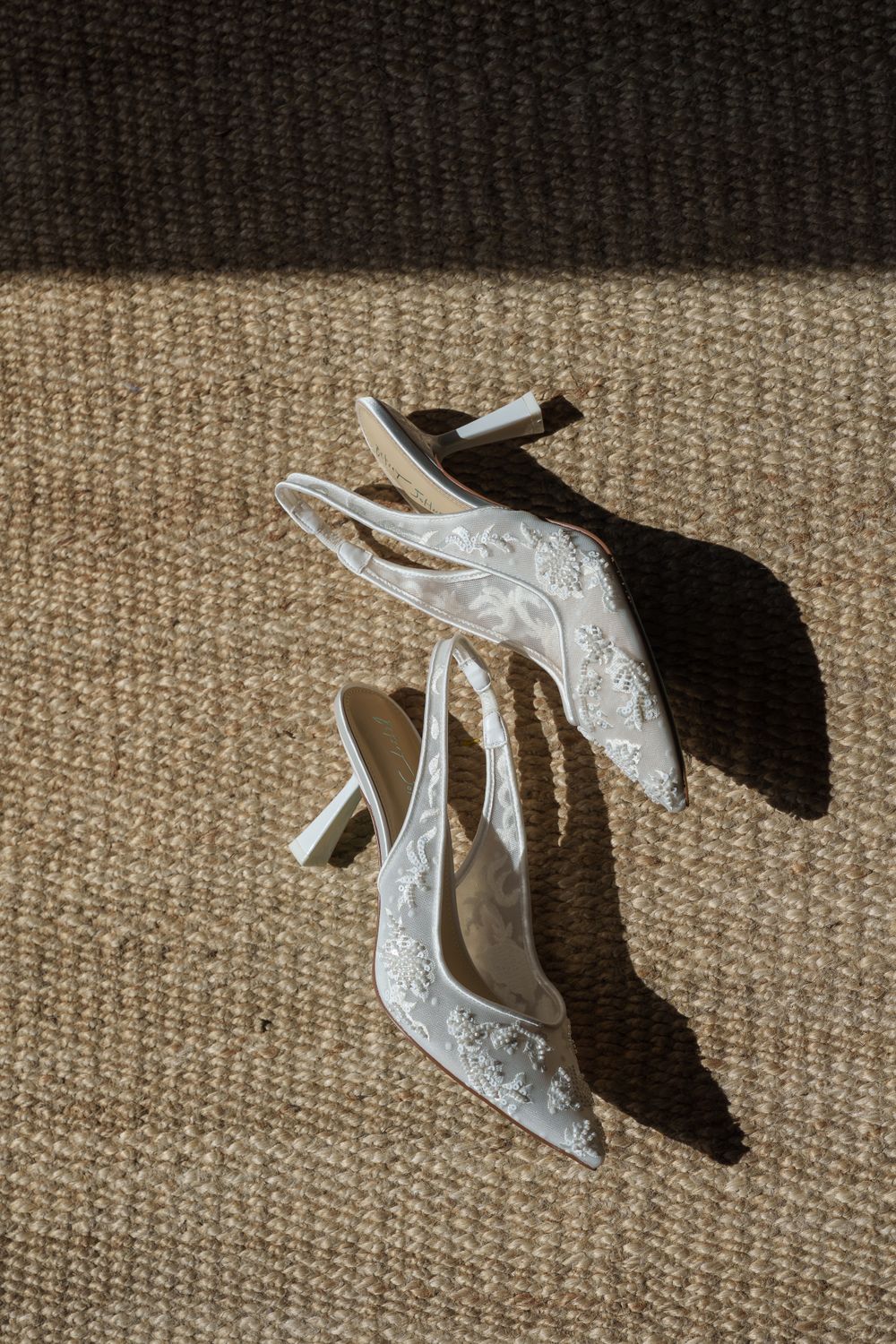 Elegant white lace wedding heels with delicate embroidery cast dramatic shadows on beige carpet.