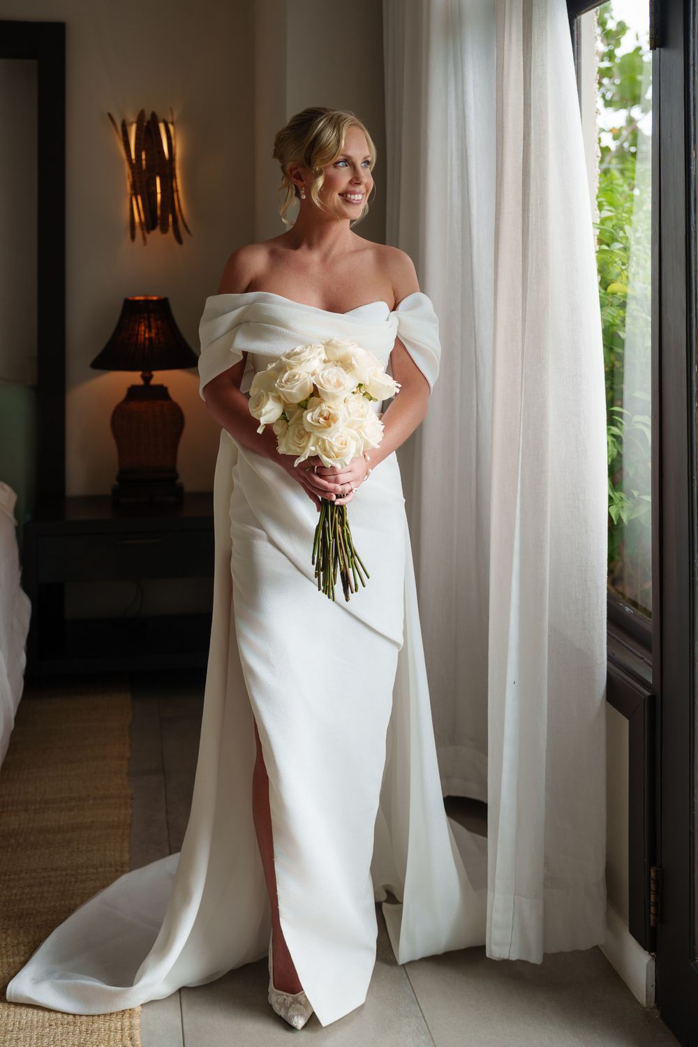 An elegant bride in a white off-shoulder wedding gown holding a cream bouquet by a window.
