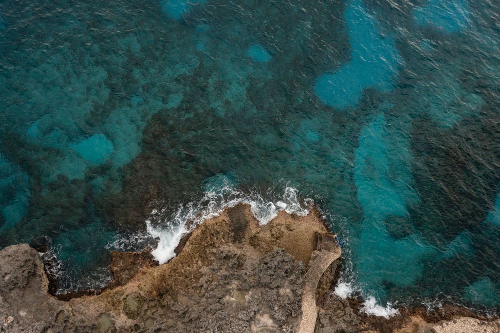 Aerial view of rocky coastline meeting turquoise ocean waters with white waves crashing against brown cliffs.