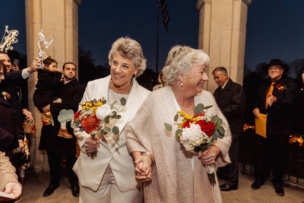 A joyful wedding couple in white outfits walk hand-in-hand holding colorful bouquets while guests celebrate behind them.