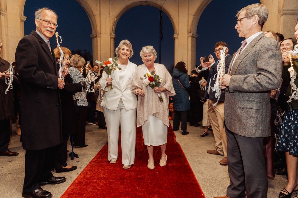 A wedding ceremony processional on a red carpet with guests looking on in an elegant arched venue.