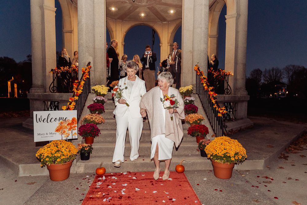 A couple walks down red carpet after their wedding ceremony with fall decorations and pumpkins in the background.