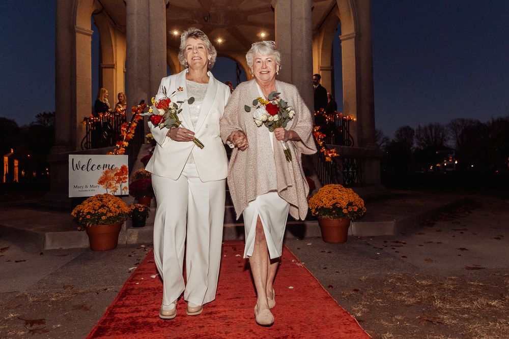 A couple poses with bouquets on a red carpet outside an elegant venue at dusk.