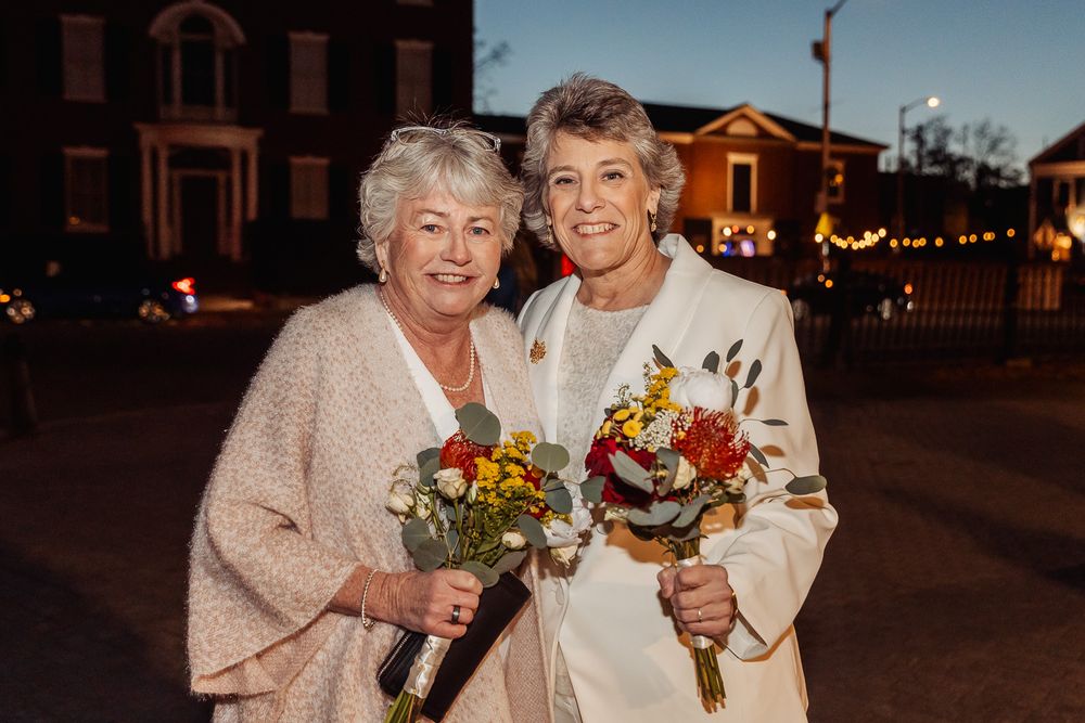 A couple holding autumn flower bouquets smiles together outside at night with buildings in the background.