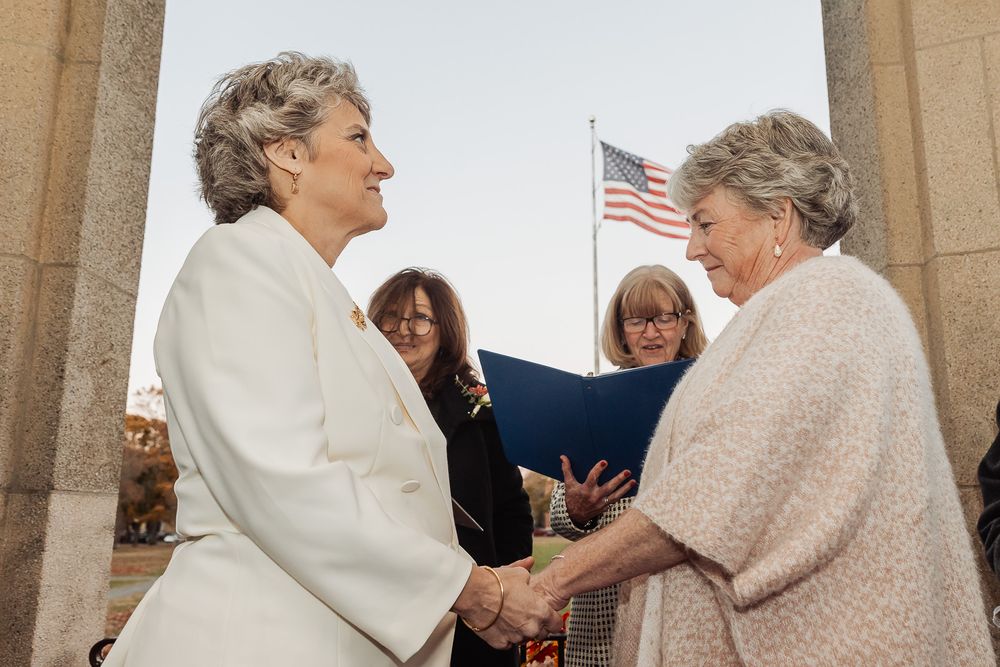 A wedding ceremony takes place under a stone archway with an American flag visible in the background.