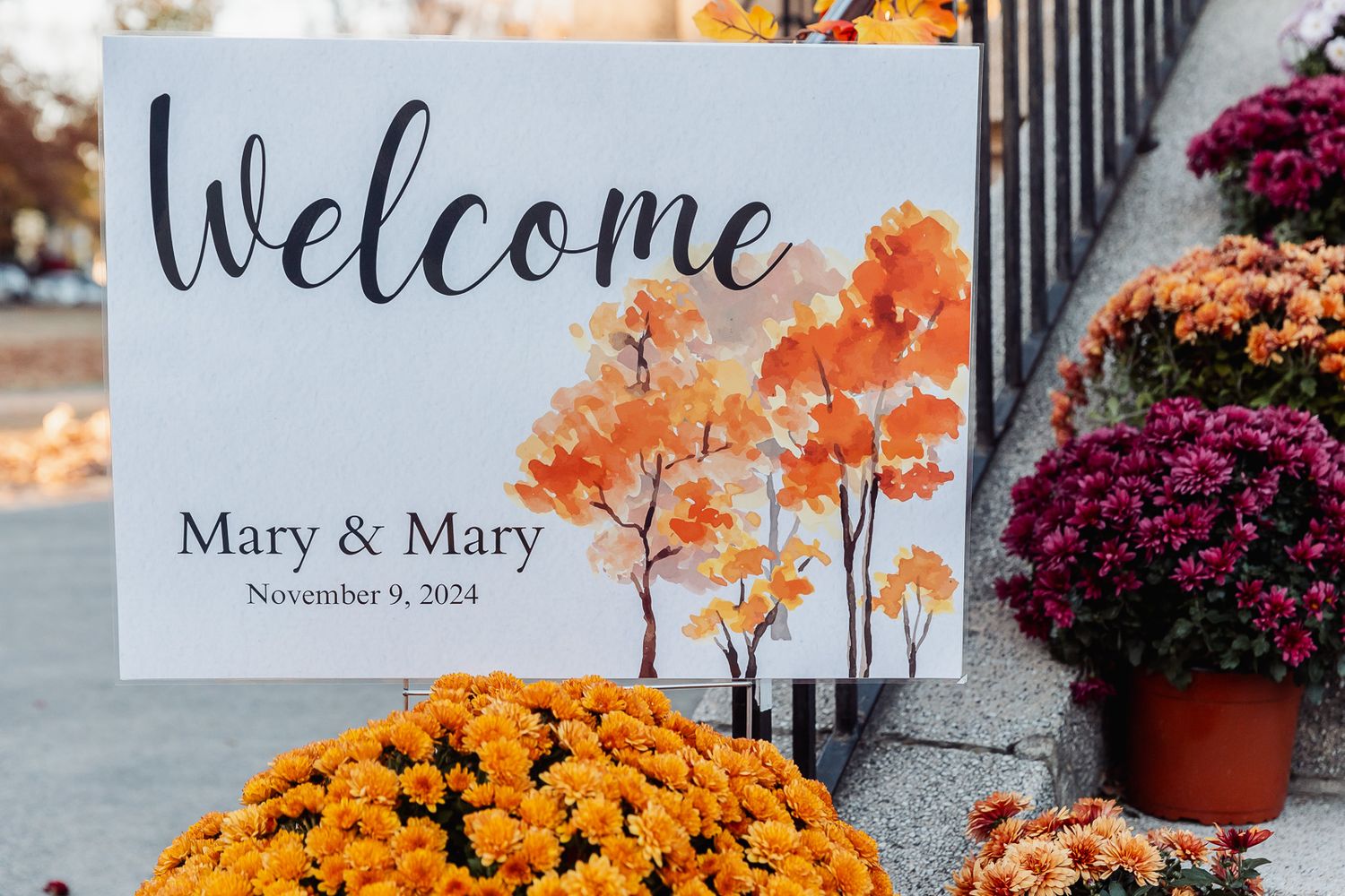 A welcome sign with autumn maple leaves sits among orange mums and pumpkins at a wedding ceremony.