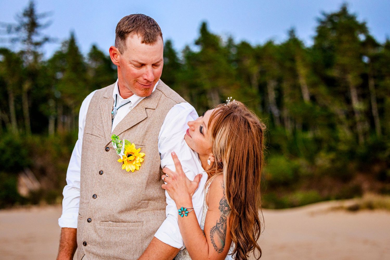 Joyful couple shares a laugh during their outdoor beach wedding ceremony with evergreen trees in background.