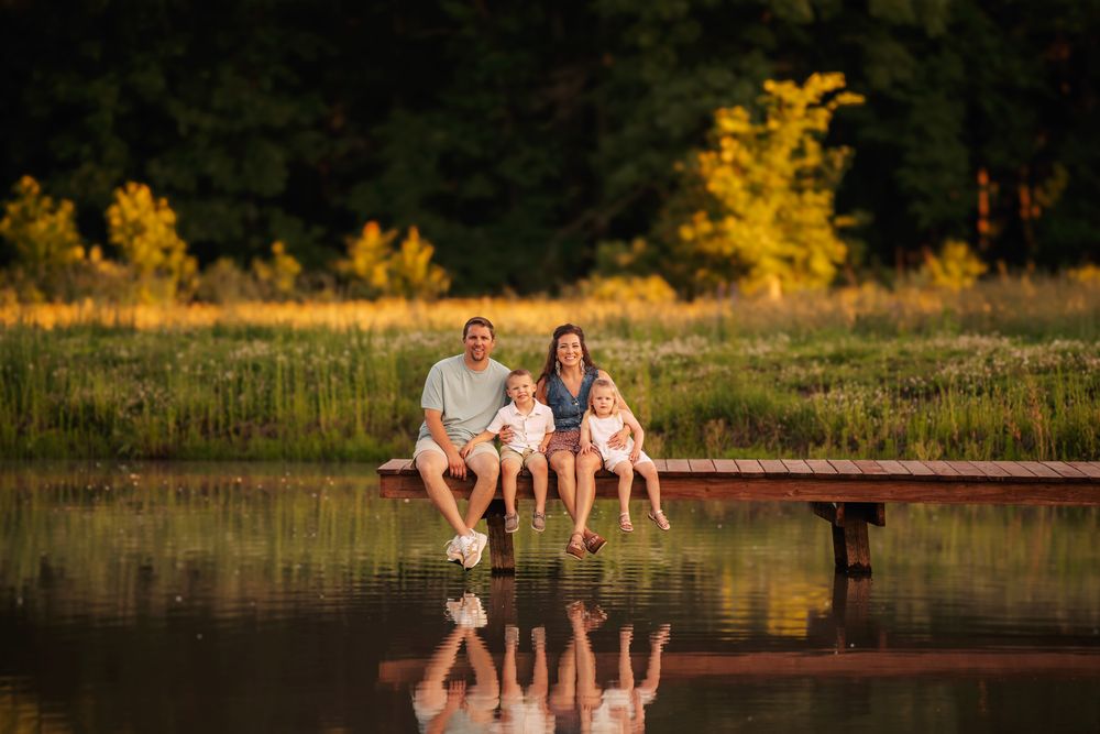 Family sits on wooden dock with their feet dangling over calm lake water at golden hour with reflections below.