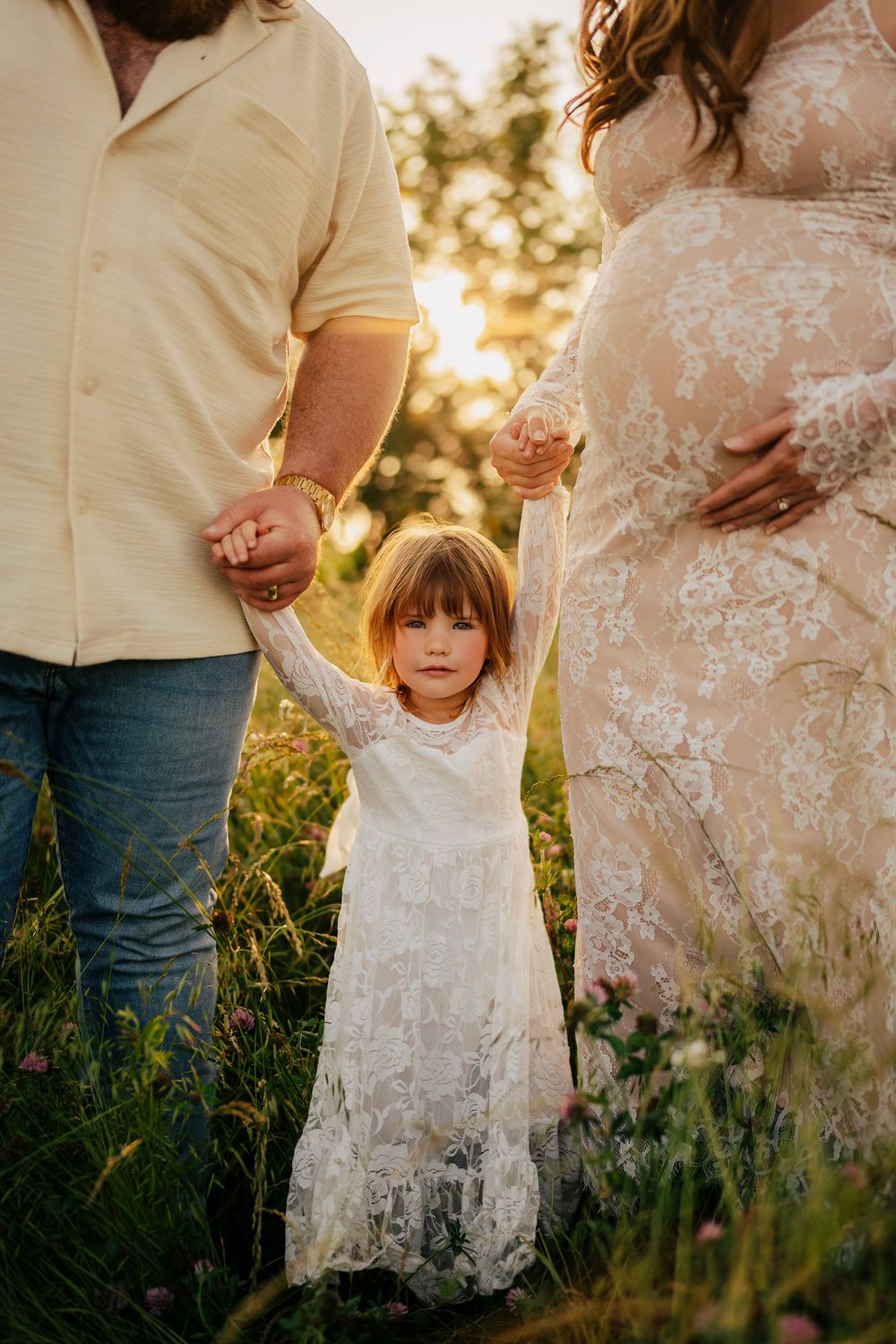 A child in a white lace dress holds hands with adults in a sunlit field at golden hour.