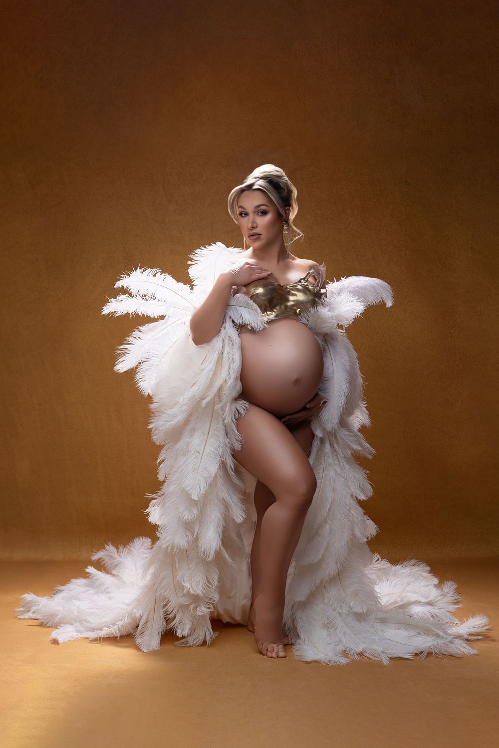 A pregnant silhouette wearing angel wings sits gracefully against a soft brown studio backdrop in artistic maternity photos.