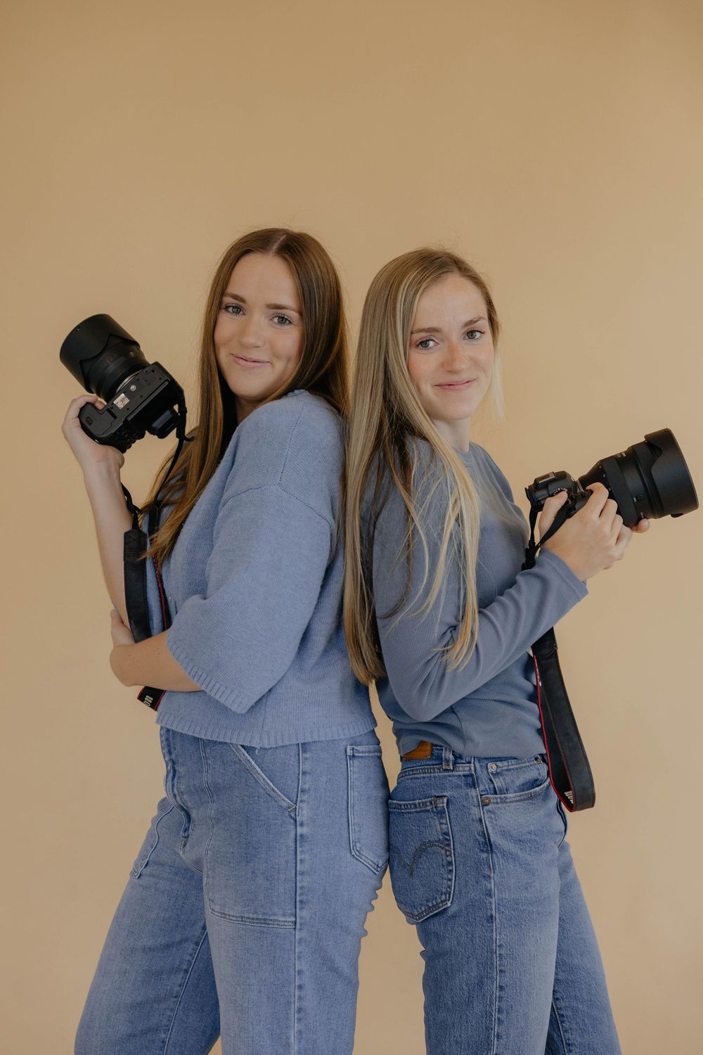 Two photographers wearing black tops and jeans stand against a white brick wall while holding professional cameras.