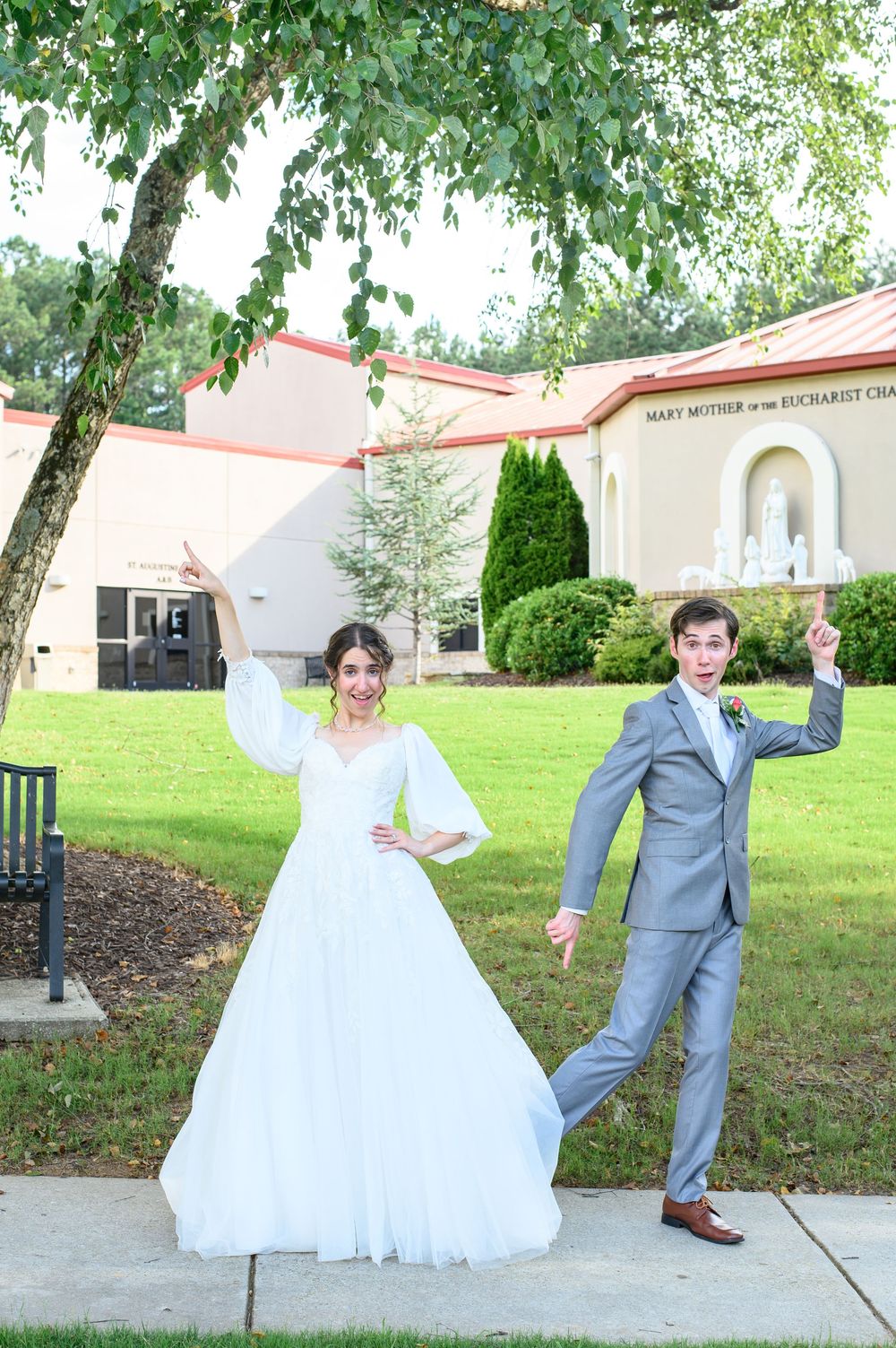 A couple poses playfully outdoors in wedding attire with the bride in a white gown and groom in gray suit.